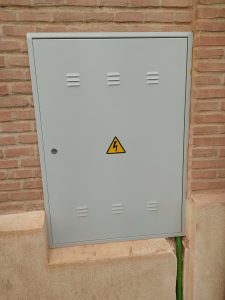 A wall with an electrical panel featuring a high-voltage warning symbol, mounted on a textured brick surface. The warning symbol prominently displays a lightning bolt, emphasizing danger.