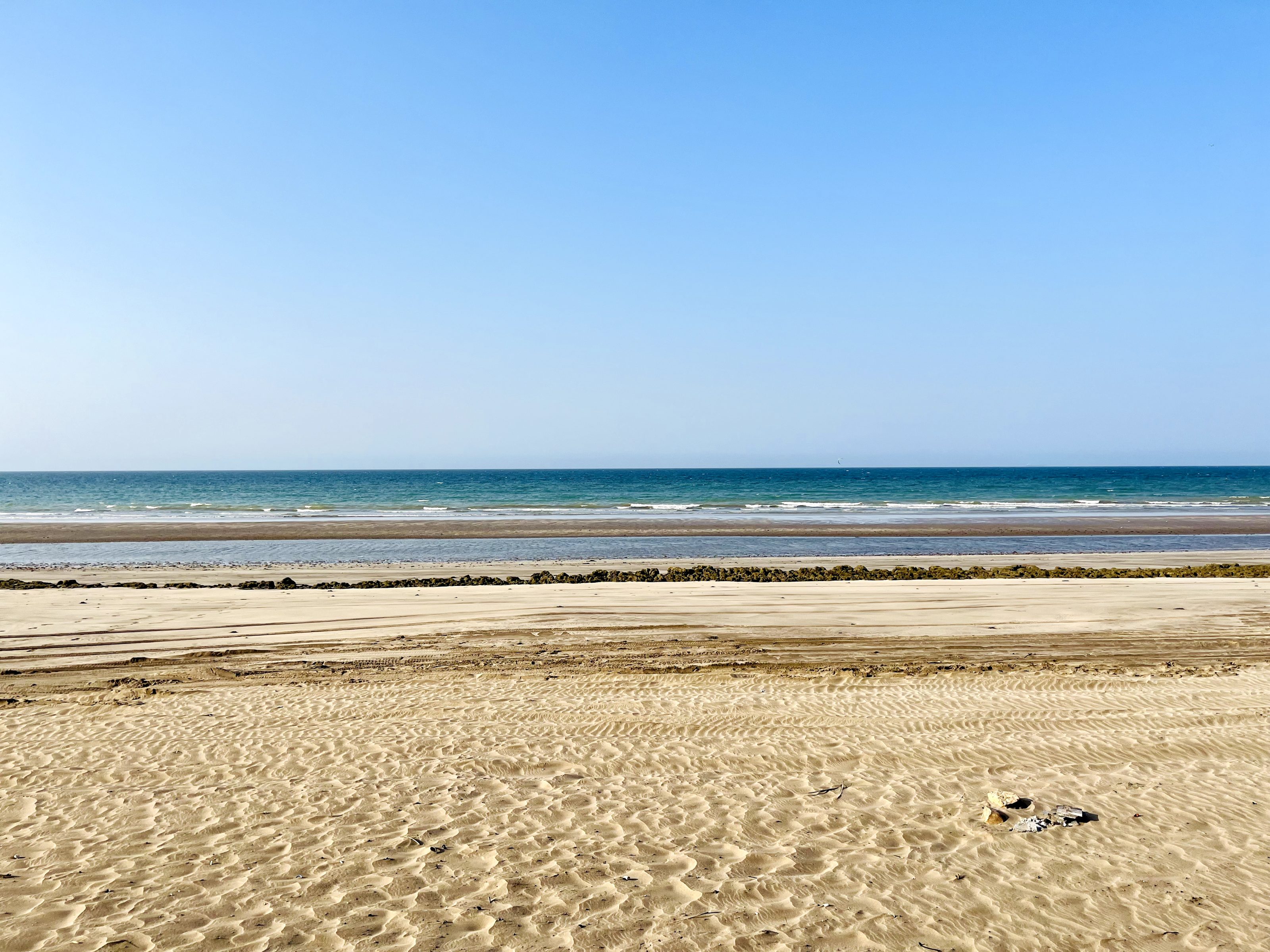 The photo shows a serene beach with golden sand stretching toward the sea. The ocean has gentle waves, and the horizon is visible under a clear blue sky. A line of rocks or seaweed marks a boundary near the water, adding texture to the landscape.