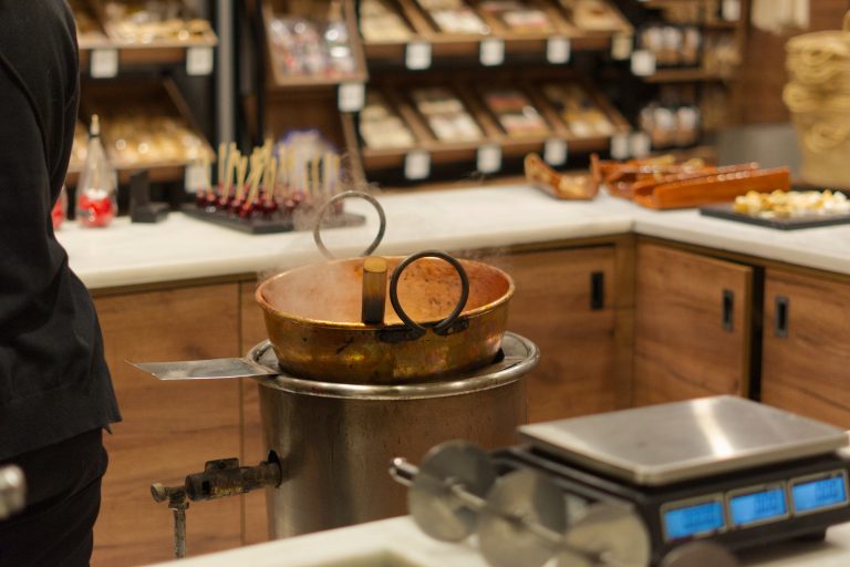 A copper pot with steam rising from it is placed on a stove in a kitchen or candy shop setting. In the background, there are shelves stocked with various goods and a tray of red candy apples.