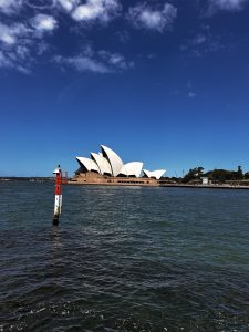 A waterfront view of the Opera House with its iconic white sail-like roofs under a clear blue sky, with a buoy visible in the water.