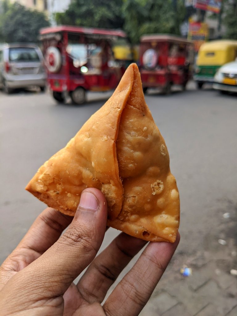 A close-up of a hand holding a crispy, golden-brown samosa with street traffic, including rickshaws and cars, visible in the blurred background.