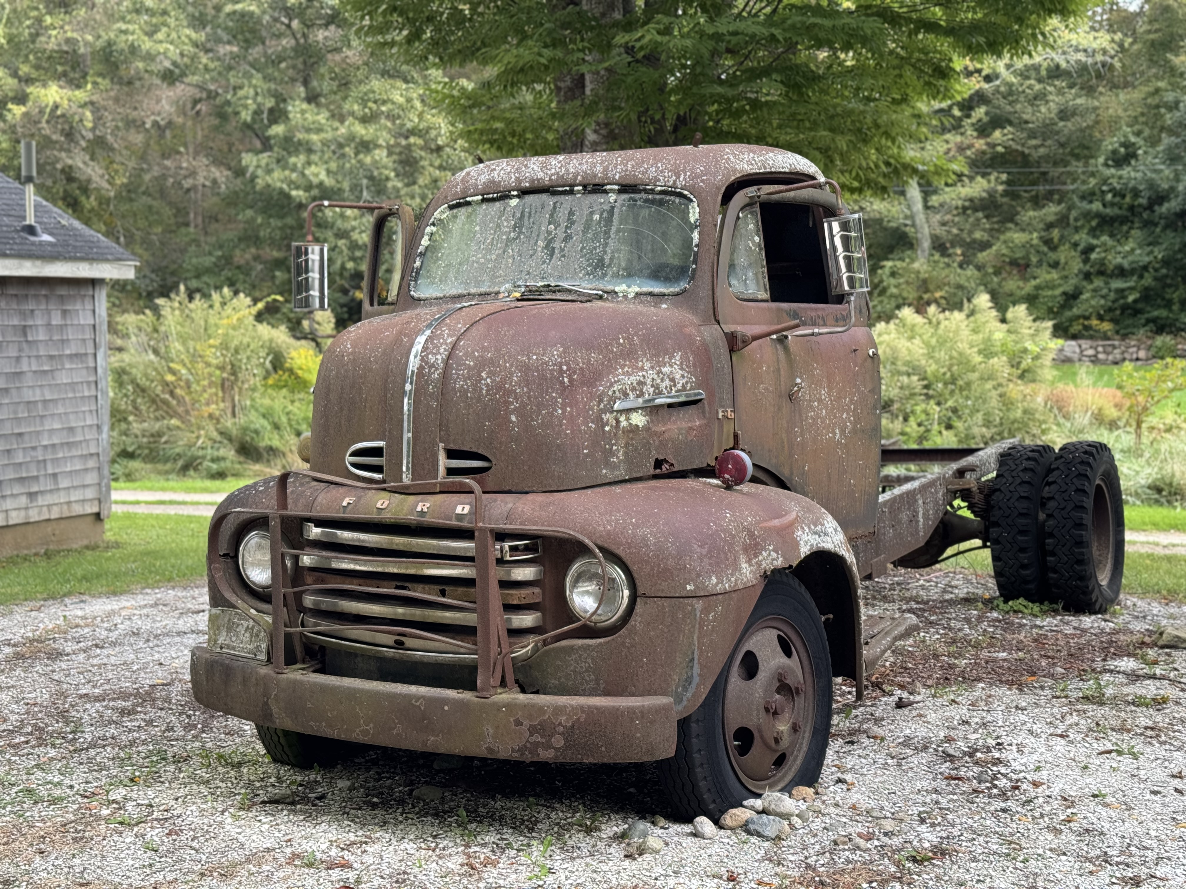 An abandoned, old, rusty Ford pickup truck parked in a gravel driveway. The pickup bed is removed.