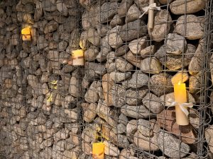 A wall of rocks held in by a metal fence is lit by electric candles that are tied to it.