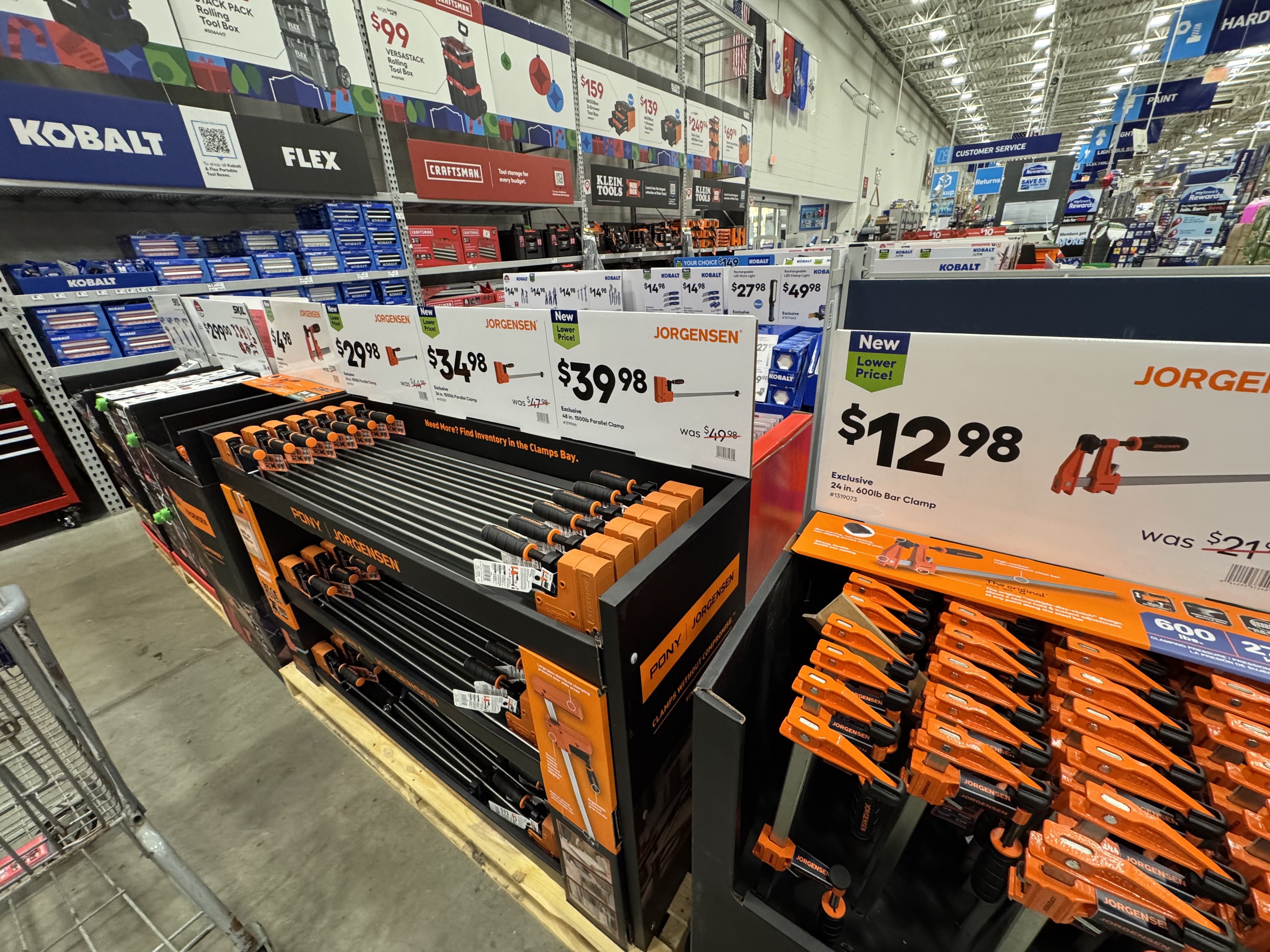 A side stack display of orange woodworking clamps at a hardware store. Shelves of tools can be seen in the background.