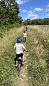 Three kids on bikes wearing helmets ride a dirt path through prairie grass through a forest preserve