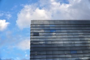 A modern glass office building with reflective windows against a backdrop of a partly cloudy blue sky.