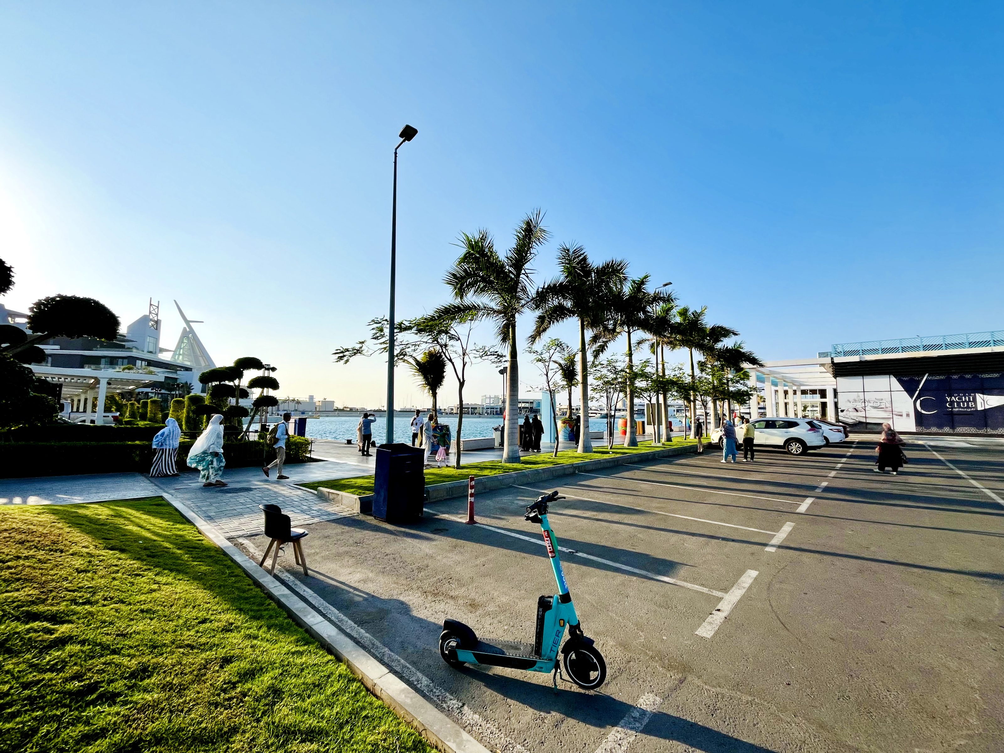 The image shows an outdoor setting with a clear blue sky, palm trees, and a waterfront in the background. A parking lot is in the foreground, where a bright turquoise e-scooter stands. People are walking near the waterfront, and a modern building with a distinct sail-shaped design is visible in the distance.