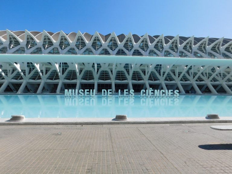 The image shows the exterior of the Museu de les Ciències, a modern architectural building with intricate white lattice structures. There is a large, reflective pool in front, and the name of the museum is spelled out in large letters above the water. Valencia, Espa?a.