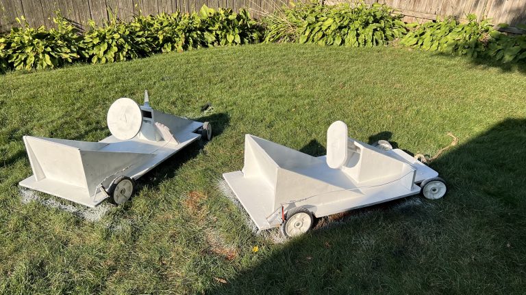 Two handmade wooden soapbox derby cards primed in white pain sitting on a yard of green grass lined with shrubs and a wooden fence