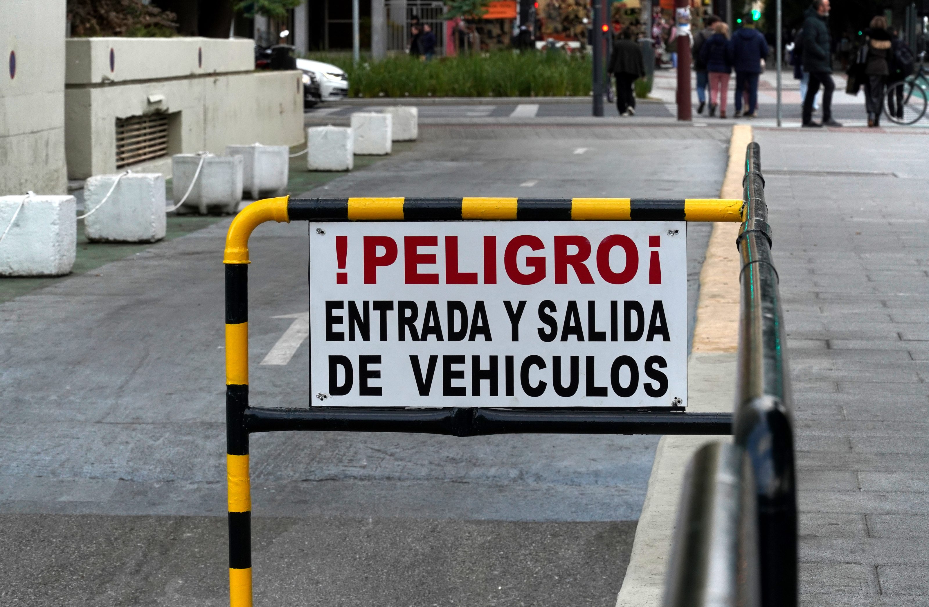 A sign on a barrier reads "¡PELIGRO! ENTRADA Y SALIDA DE VEHÍCULOS," warning about vehicle entry and exit. The scene is on a city street with people walking in the background.