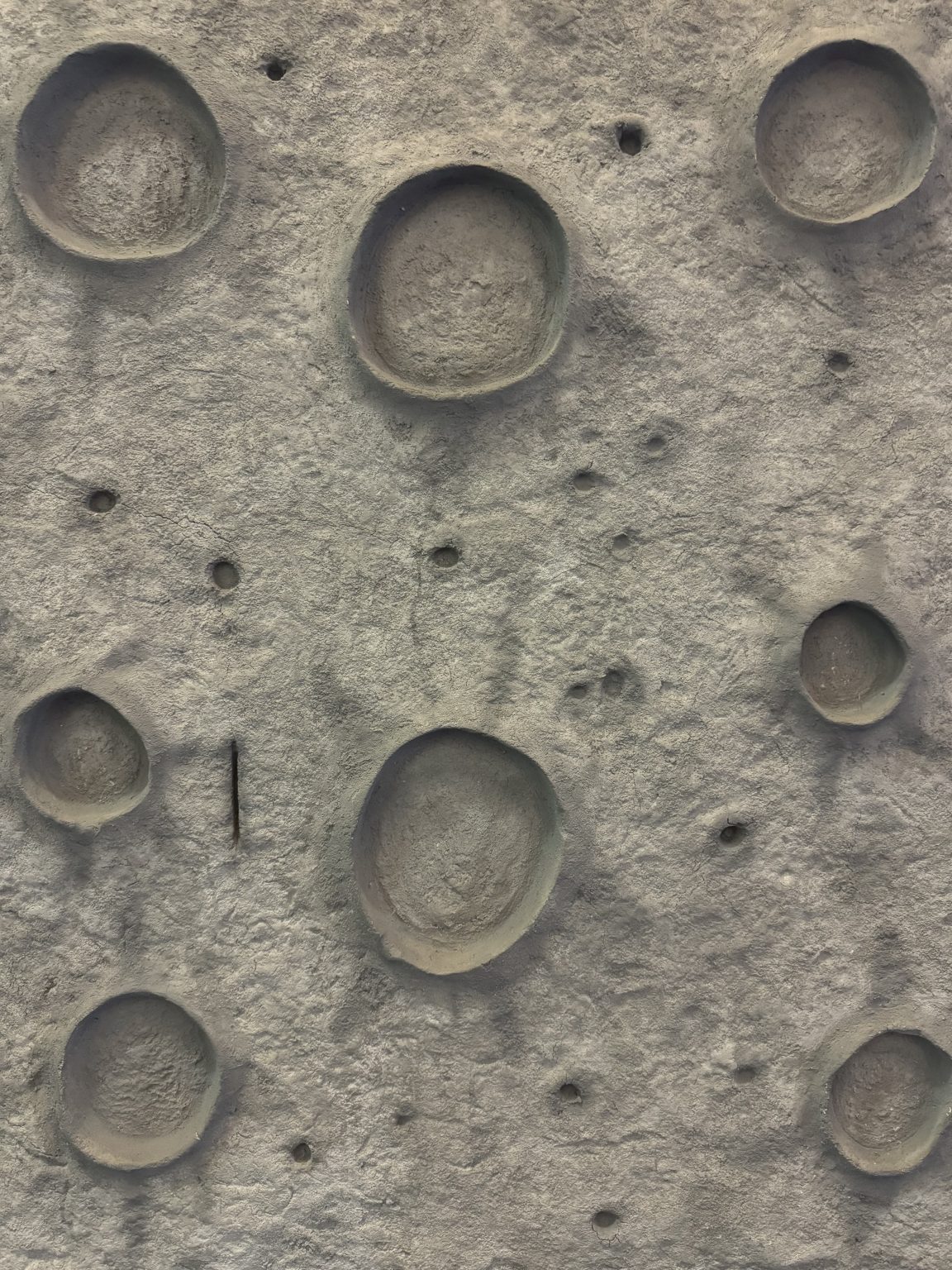 A close up of a grey climbing wall at a playground. The wall is textured like the moon’s surface with craters and rough areas.