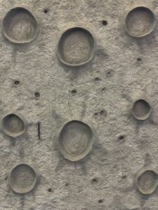 A close up of a grey climbing wall at a playground. The wall is textured like the moon's surface with craters and rough areas.