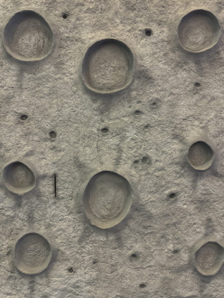 A close up of a grey climbing wall at a playground. The wall is textured like the moon’s surface with craters and rough areas.