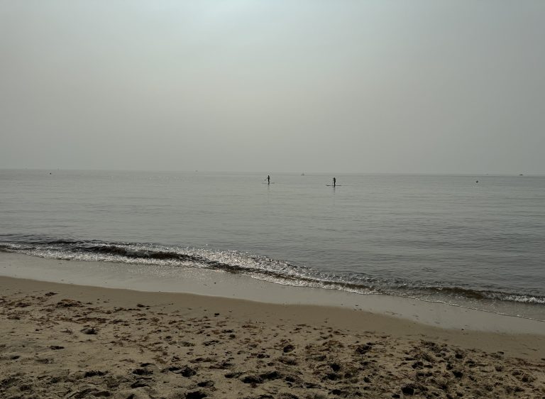 Two people paddle boarding in the ocean as seen from the beach on a hazy summer morning. Waves and disturbed sand are in the foreground.