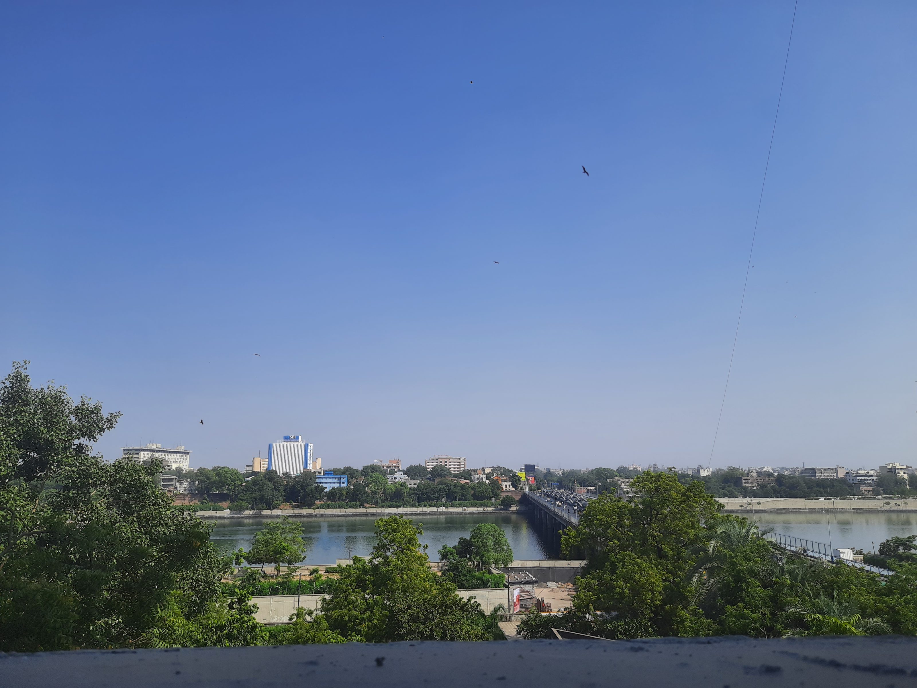 A scenic daytime view of a cityscape with a clear blue sky, a bridge spanning a river, lush greenery in the foreground, and various buildings in the background. Birds can be seen flying in the sky.