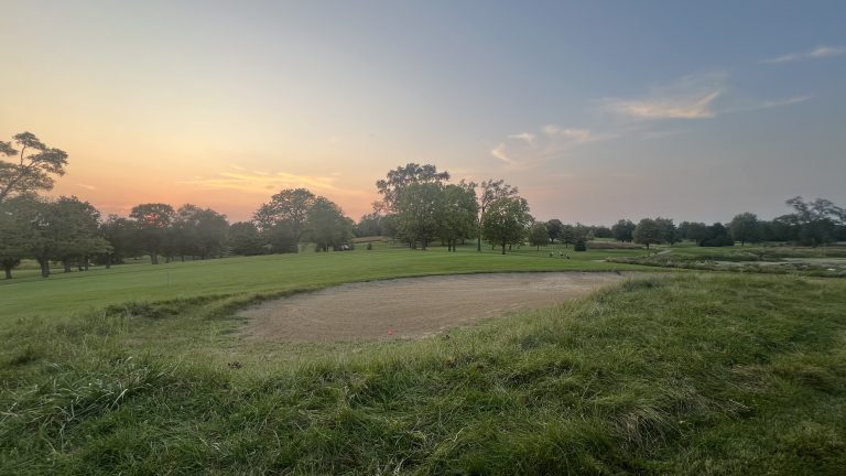 A pink golf ball sits in a sand trap as a group of golfers crests a hill in the distance at twilight and a sunset in the distance