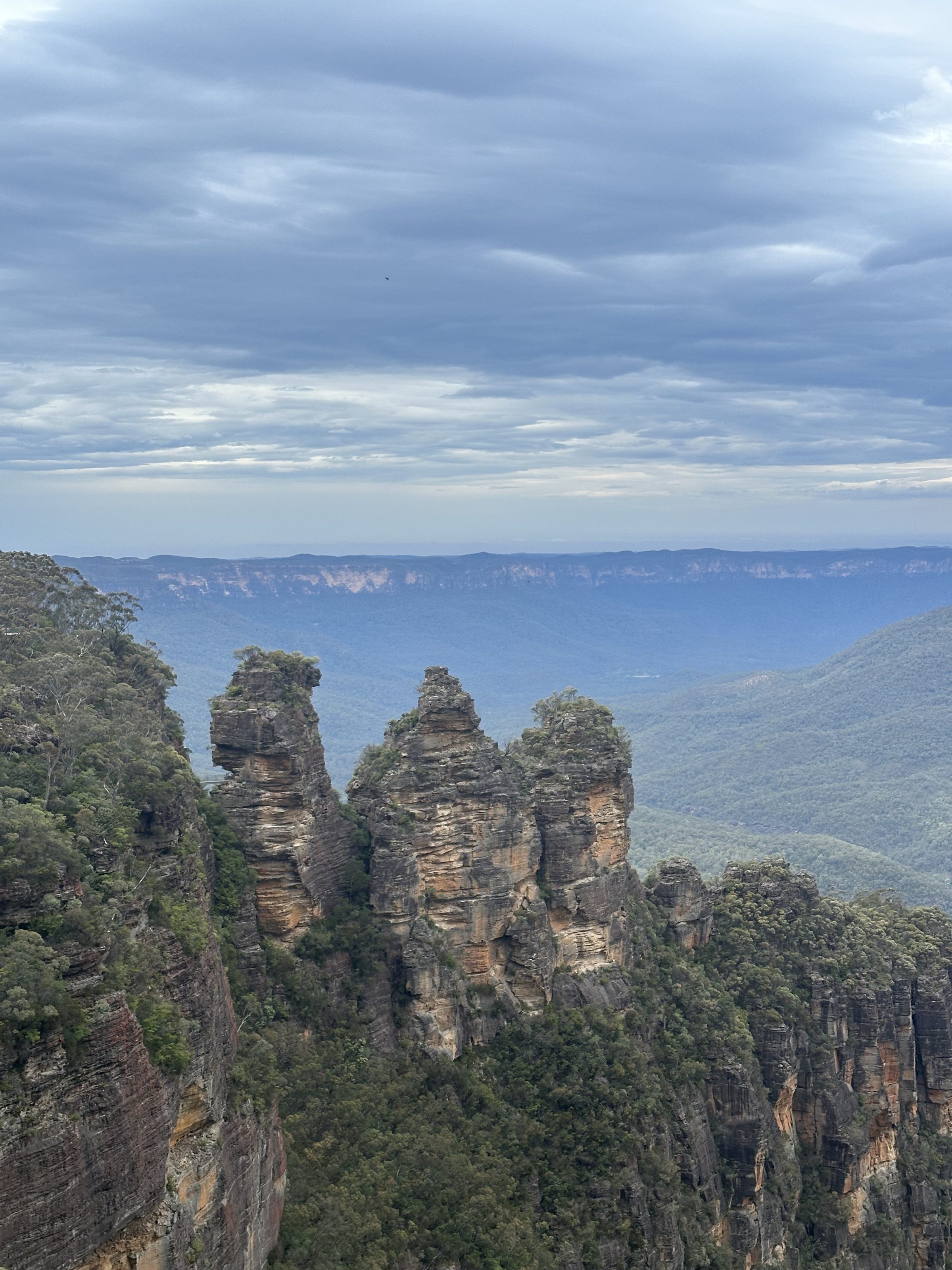 A scenic view of the Three Sisters rock formation surrounded by lush greenery in the Blue Mountains, under a cloudy sky.