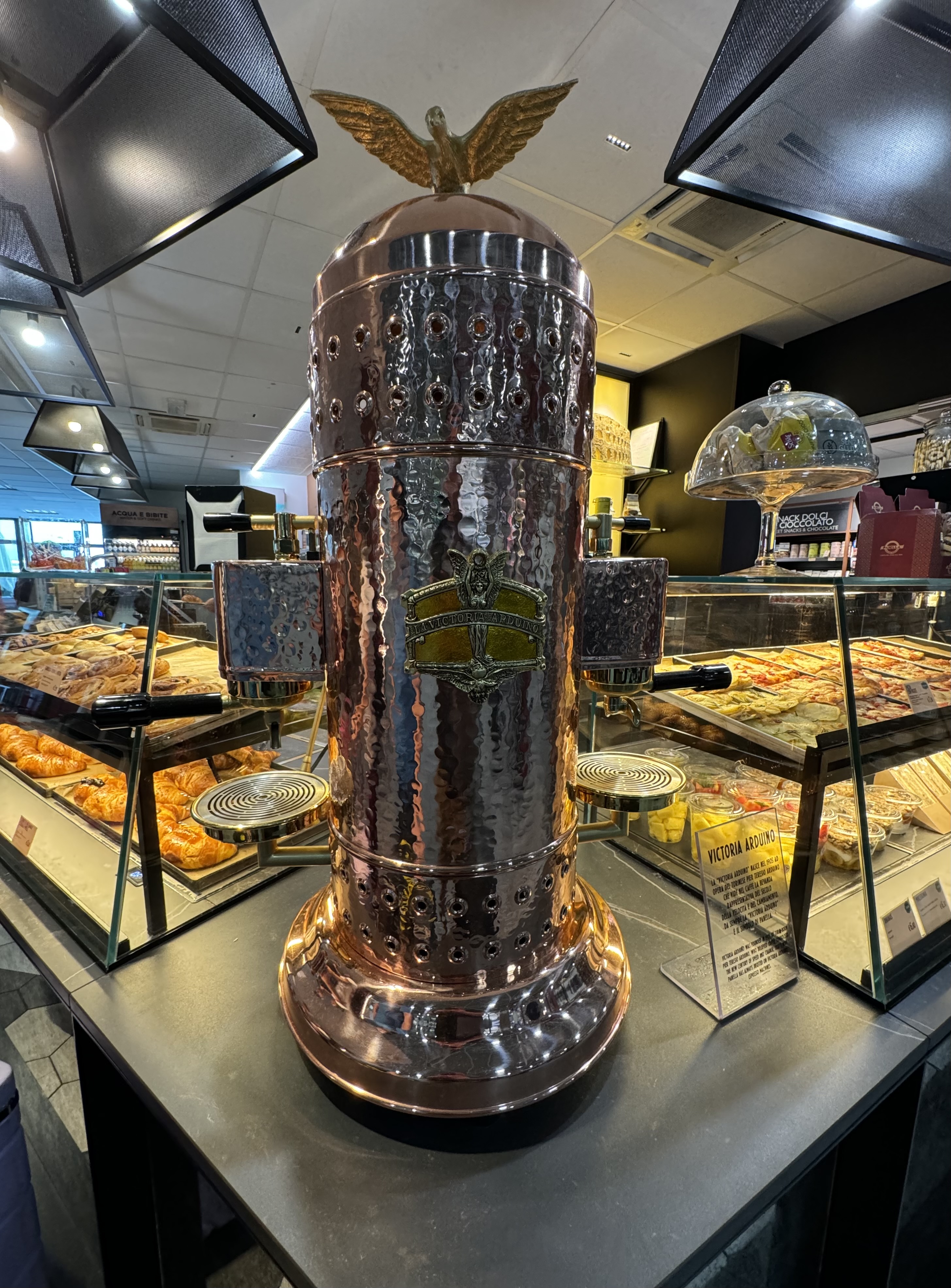 A large copper la victoria arduino coffee maker on display at a coffee shop in the Torino Airport.