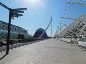 A modern architectural scene featuring the Ciudad de las artes y de las ciencias in Valencia, Spain. The image shows sleek, futuristic structures with a bright blue sky above. A directional sign in the foreground points to various attractions within the complex. T