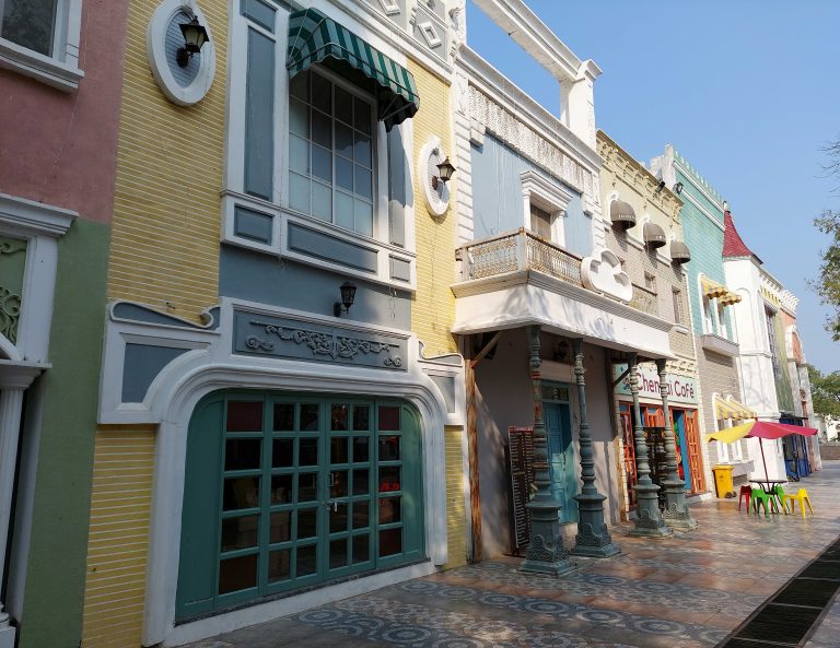 A bright and colorful street scene featuring a row of intricately designed buildings with vintage-style architecture. The street is paved with patterned tiles, and the foreground includes outdoor seating with vibrant red, yellow, and green chairs under matching umbrellas.