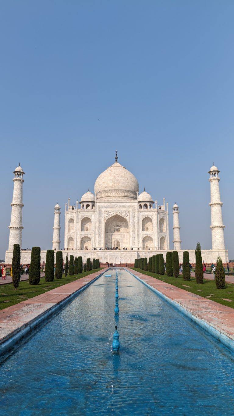 A distant view of the Taj Mahal, with its iconic white marble domes and four minarets, set against a clear blue sky. In the foreground, a long reflective pool is lined with neatly trimmed hedges, creating a symmetrical pathway leading up to the monument.