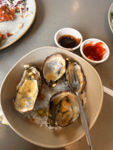 A bowl of oysters on a bed of salt is served with a small fork. Two small cups of sauce, one dark and one red, are on the side. 