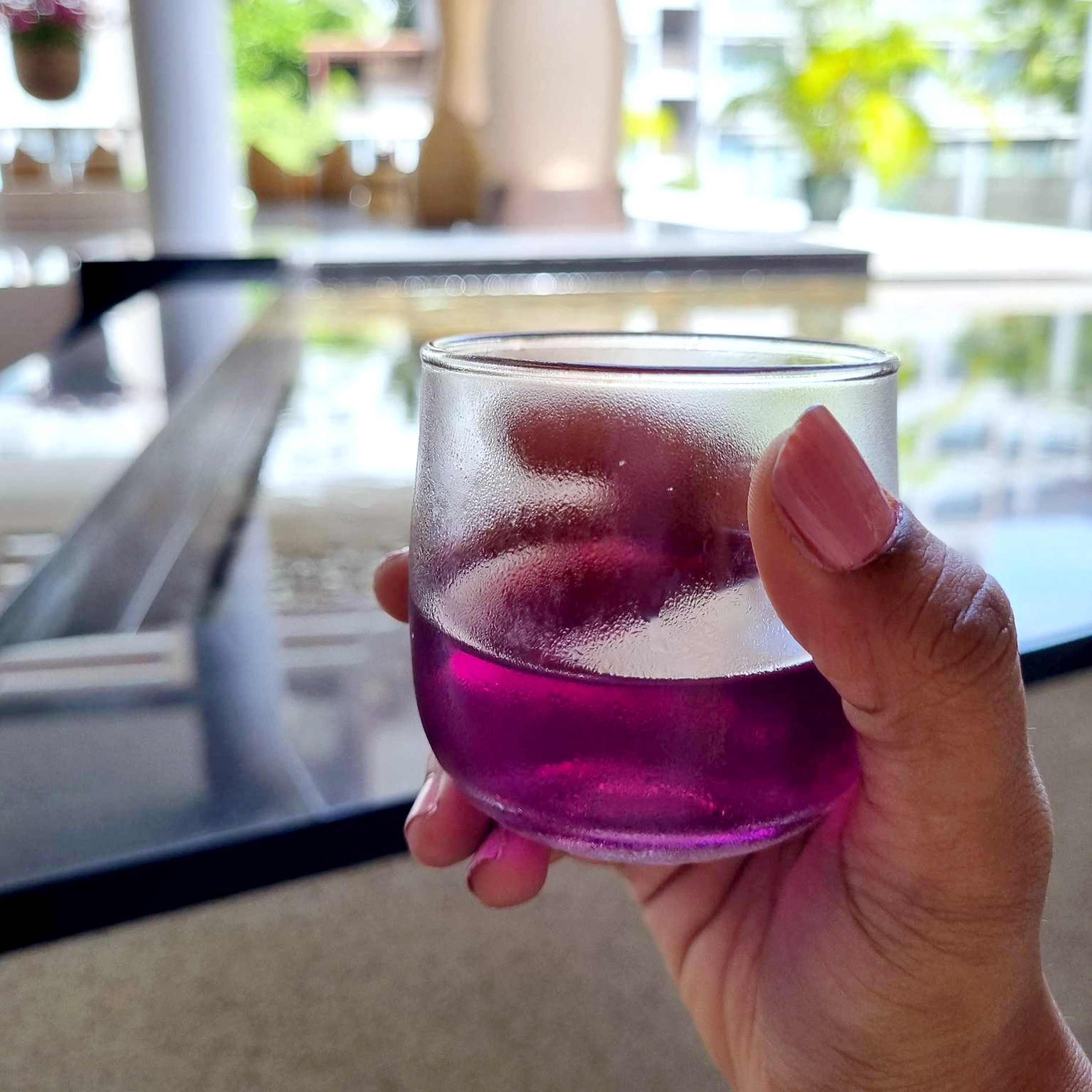 A person's hand holding a glass of purple liquid. The background shows an outdoor setting with blurred green plants and structures, and a reflective surface nearby.