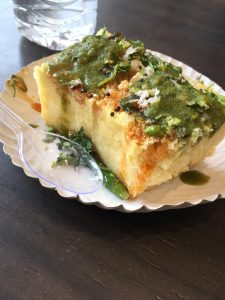 A serving of dhokla on a paper plate, topped with green chutney, coriander leaves, and mustard seeds. A plastic spoon is placed beside the dhokla, and there's a blurred glass of water in the background