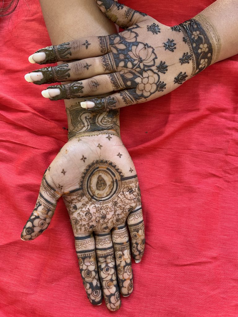 A pair of hands with elaborate mehndi designs, featuring floral and intricate patterns, resting on a red cloth. The designs cover the fingers, palms, and extend up the wrist, highlighting traditional henna art.