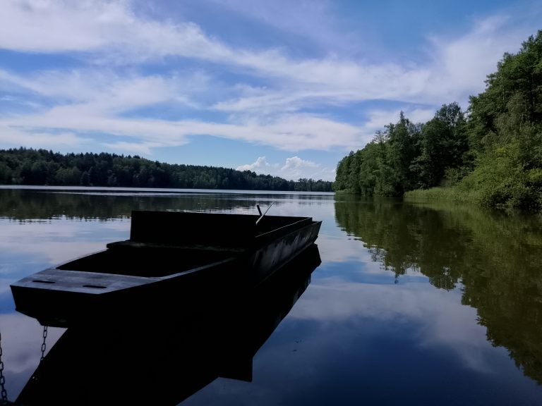 A quiet lake surrounded by a dense green forest under a partly cloudy blue sky. A wooden boat is anchored in the foreground, casting a silhouette on the calm water, which reflects the sky and trees. The serene scene conveys a peaceful, natural atmosphere.