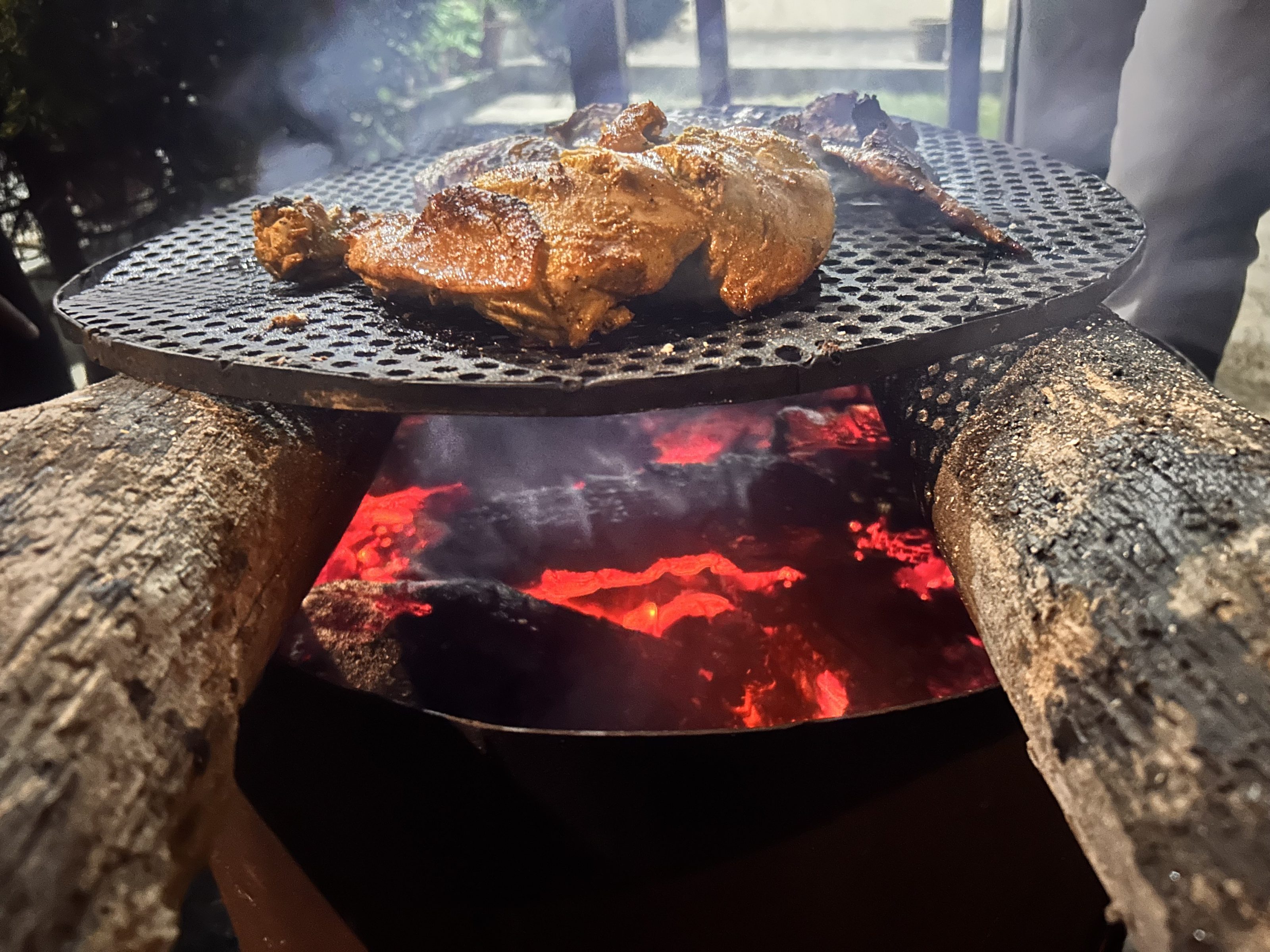 Chicken meat grilling on a metal grate over glowing red coals, supported by logs, with smoke rising from the fire.