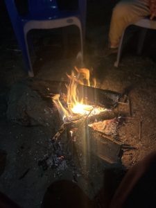 A small campfire burning on the ground, surrounded by logs, with two chairs partially visible in the background. The scene is dimly lit, highlighting the warm glow of the fire.