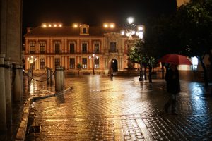 A nighttime scene of a historic building with a lit facade in the background, featuring wet cobblestone streets reflecting the lights. A person with a red umbrella walks in the foreground