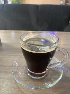  clear glass cup of black coffee on a saucer, placed on a wooden table with a spoon next to it. The background shows a soft-focus view of a dark upholstered couch.