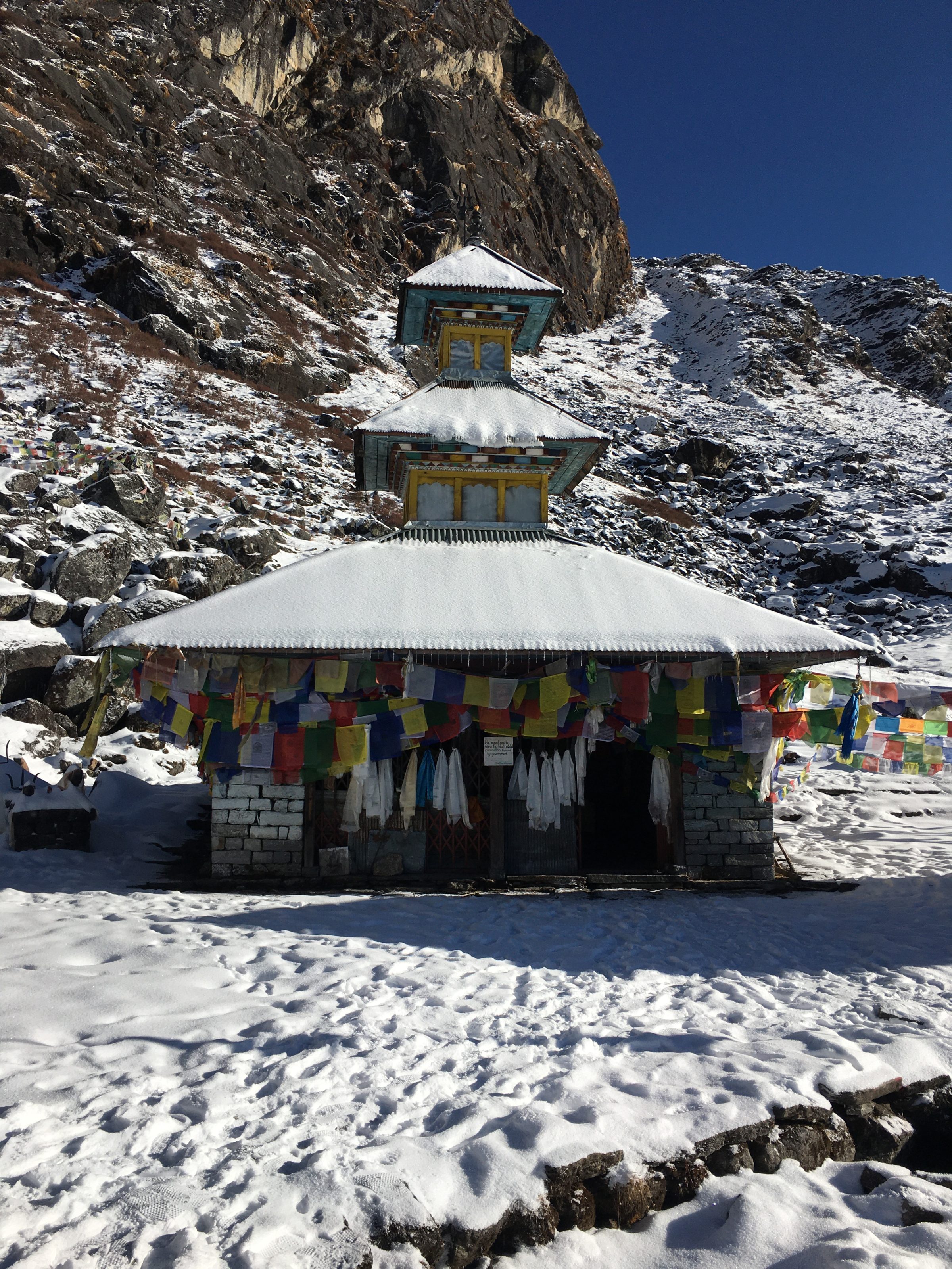 A small, snow-covered building with colorful prayer flags in a mountainous landscape. The structure has a tiered roof and is set against a backdrop of rugged, rocky terrain under a clear blue sky.