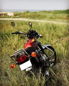 A red motorcycle parked in tall grass, with a scenic view of a distant road and greenery in the background.