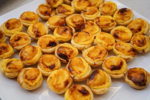 A close-up view of several freshly baked Portuguese egg tarts (pastéis de nata) arranged on a white plate. The tarts have a crispy, flaky pastry crust and a golden-yellow custard filling with a slightly browned top.