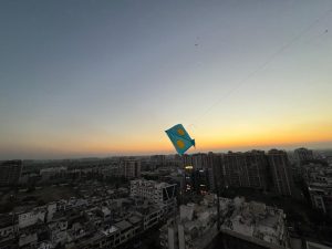 A blue kite with yellow dots soars high in the sky against a backdrop of a colorful sunset and cityscape.