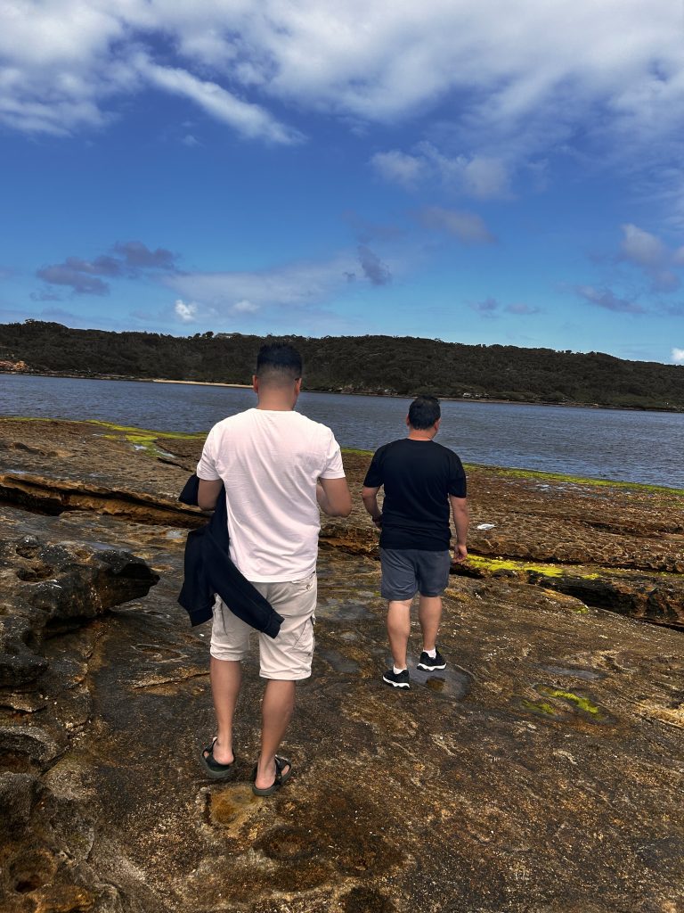 Two people standing on rocky terrain near a body of water, with a distant treeline and a partly cloudy sky in the background.