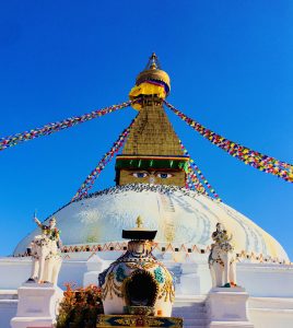 A large white Boudha stupa with a golden spire, adorned with colorful prayer flags stretching from the top. 