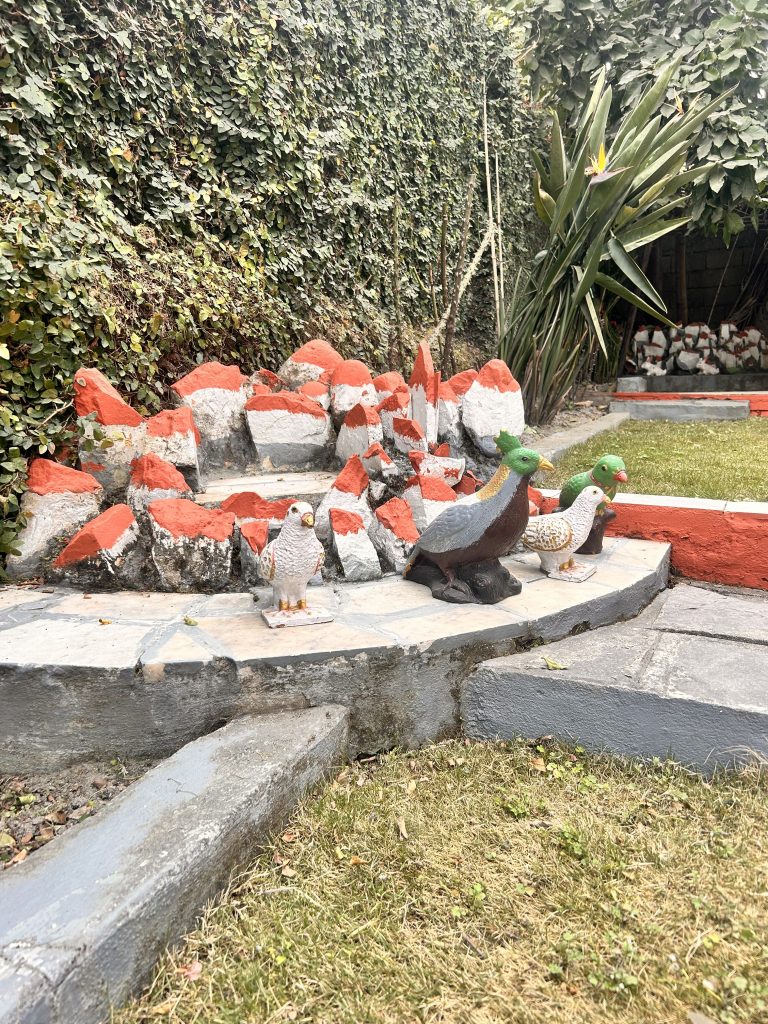 Garden scene featuring colorful painted stone sculptures of birds positioned on a stone platform. The background includes a lush hedge wall and some large green plants, with neatly trimmed grass in the foreground.