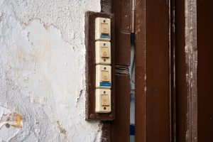 A set of four vintage doorbells mounted on a textured wall next to a brown doorframe. The wall has peeling white paint, and the buttons are labeled with small icons and text.