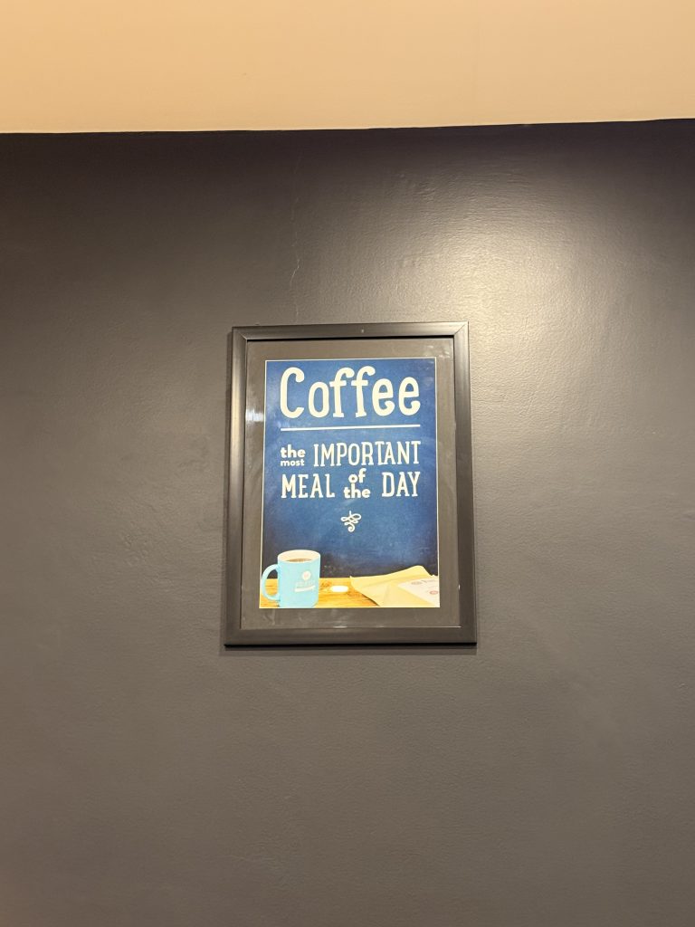 A framed poster hanging on a dark wall. The poster features a retro-style design with a blue background and an illustration of a coffee cup next to an envelope. The text in large white letters reads, “Coffee the most important MEAL of the DAY.”