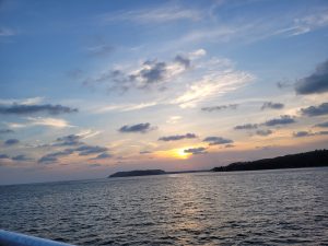 A serene sunset over the ocean with a clear sky featuring scattered clouds, the horizon showing a silhouette of distant land, and the sea reflecting the colors of the sunset.