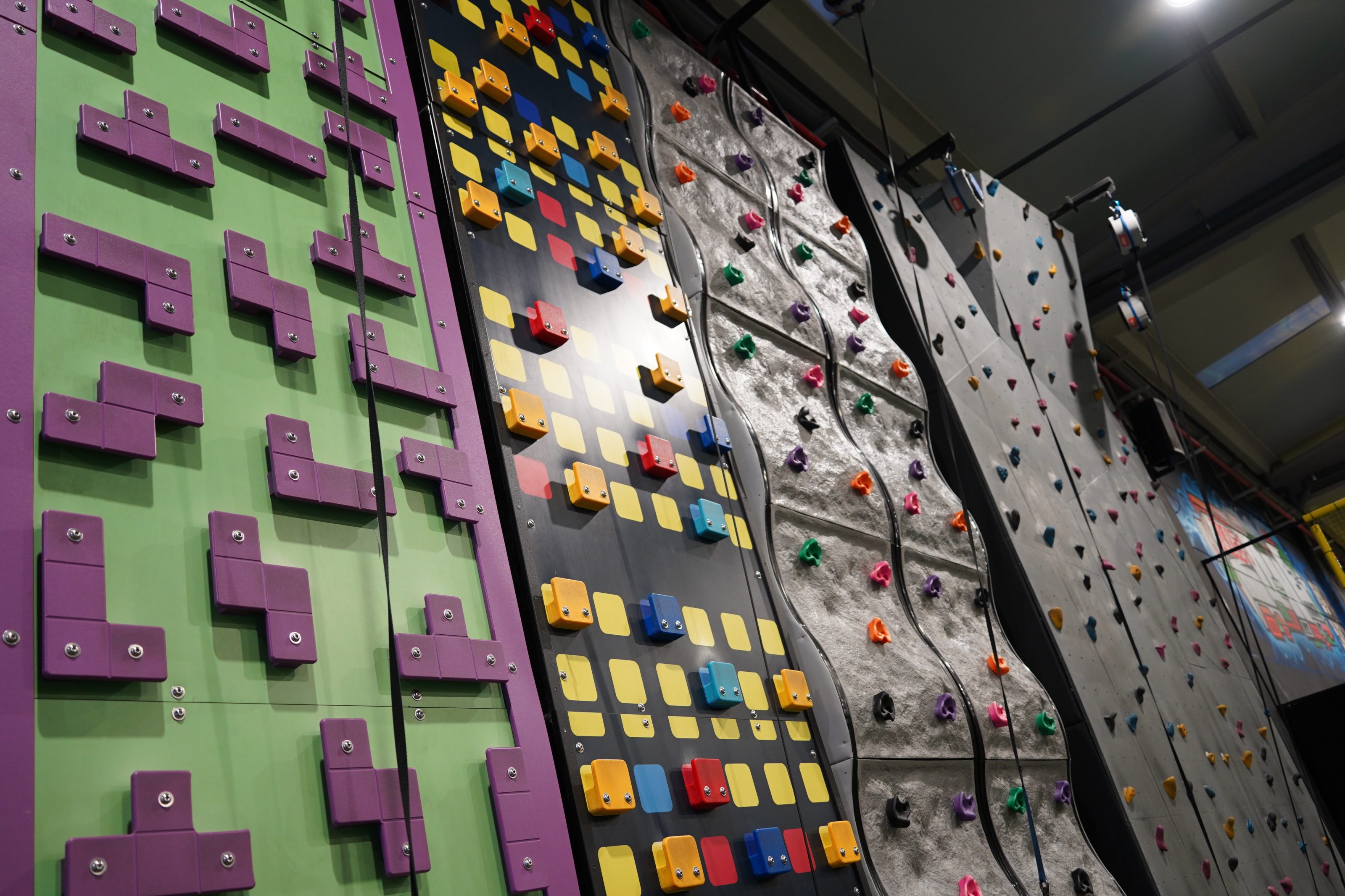 Indoor climbing facility featuring multiple climbing walls with various designs and difficulty levels. The walls include colorful, geometric holds and textures, offering diverse climbing challenges