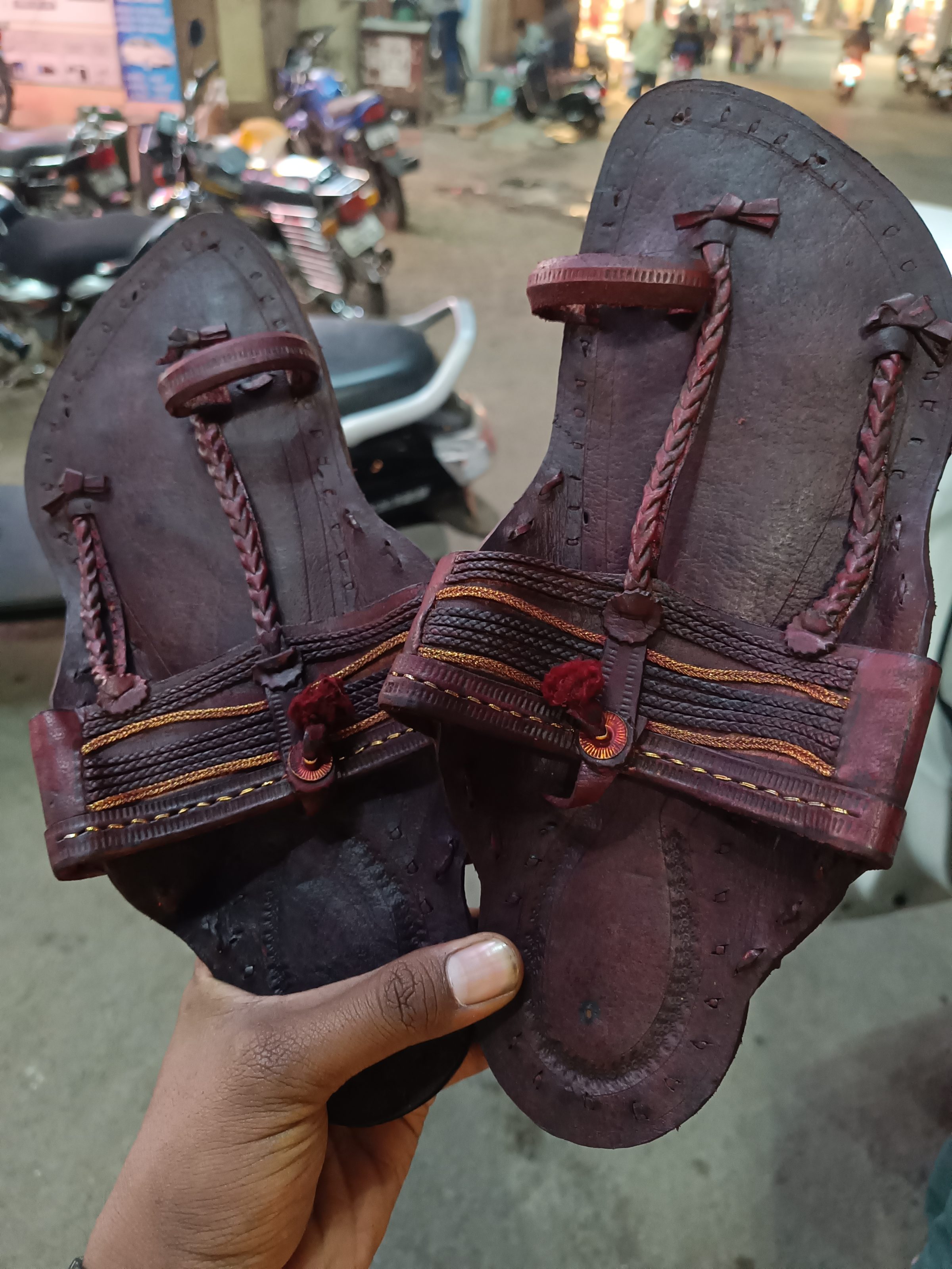 A person holding a pair of handcrafted leather sandals (Known as Kolhapuri Paytan or Kolhapuri Chappal) on a street with motorcycles and shops in the background.