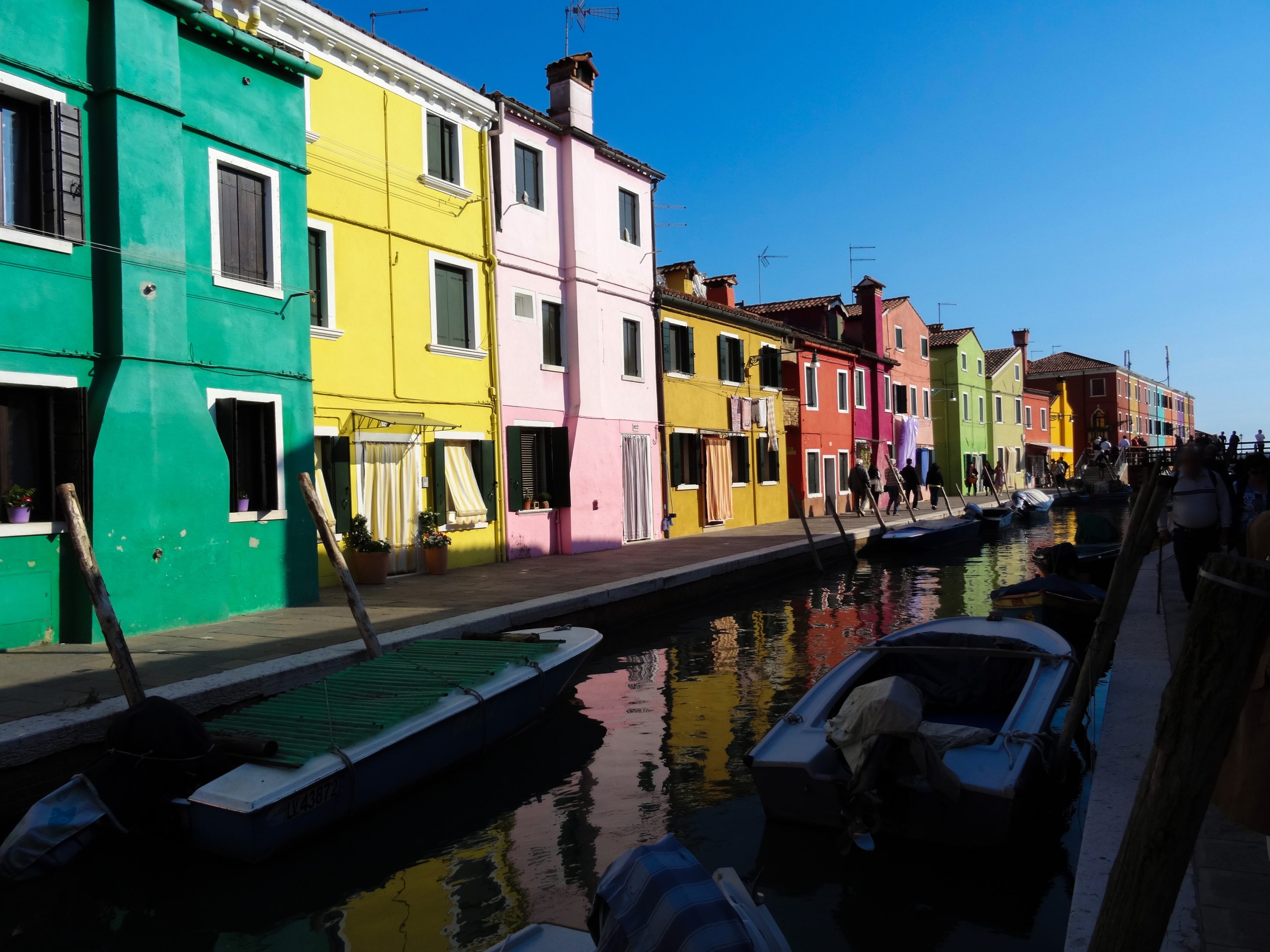 A picturesque view of a canal lined with vibrant, colorful buildings in shades of green, yellow, pink, red, and orange under a clear blue sky. Small boats are moored along the canal, and several people are walking along the waterfront.