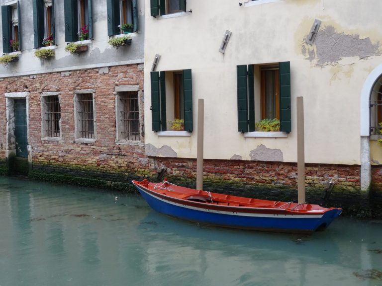 A small blue and red boat is floating on a canal in Venice, Italy, next to a building with green shuttered windows and weathered brick walls. The water appears calm, and there are potted plants on the windowsills.