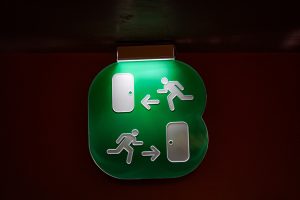 Illuminated green emergency exit sign with two directional running person icons and arrows pointing towards doors, indicating escape routes.