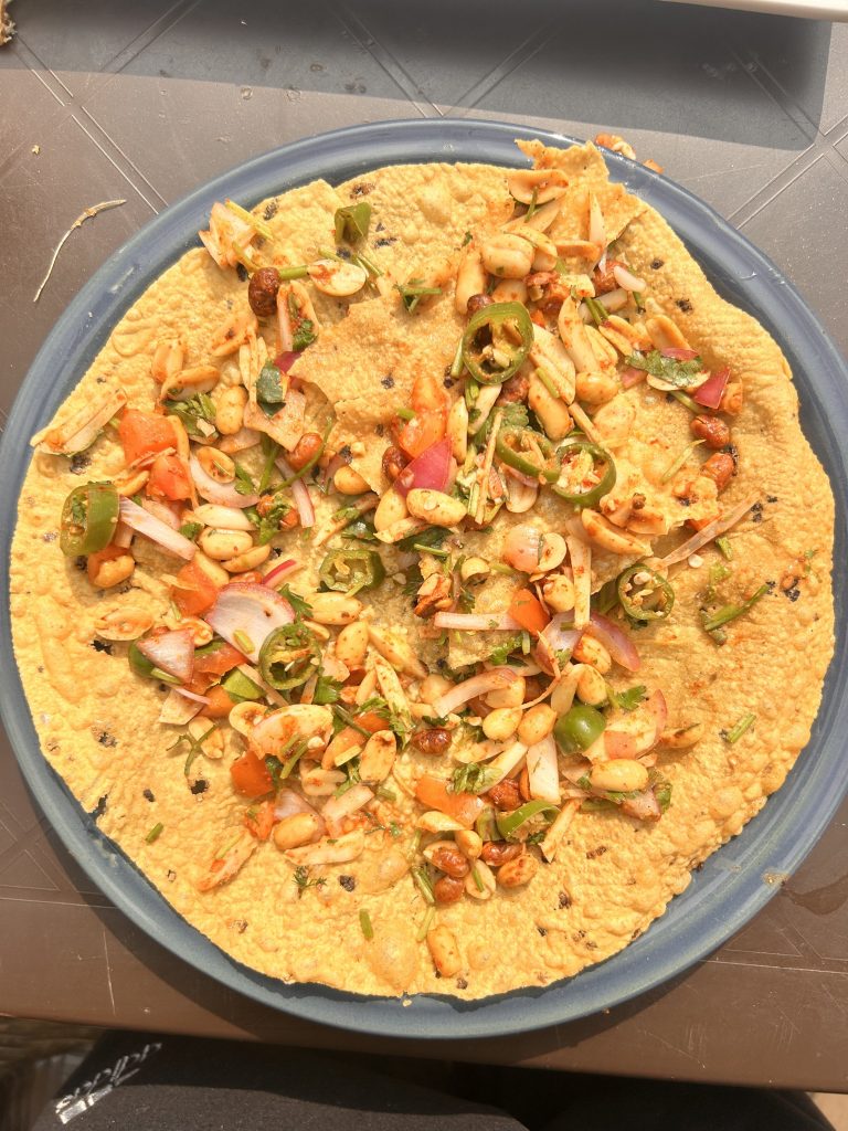 A close-up of a large, round cracker topped with a mix of chopped onions, tomatoes, greens, peanuts, and sliced green chilies, garnished with herbs and spices, placed on a blue plate.